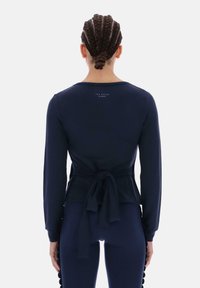 Granatowa bluzka z długim rękawem z przewiązanym w talii wiązaniem i miękkiego materiału. Posiada zaokrąglony dekolt i branding na plecach. W zestawie z dopasowanymi spodniami.