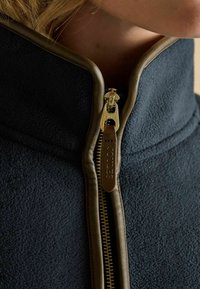 Námornícka fleecová bunda s vysokým golierom, so zlatými zipsami a koženkovými detailmi. Detailný záber ukazujúci textúru látky a detaily zipsu.