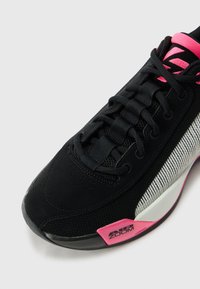 Chaussure de sport noire en matériaux mesh et daim, avec des accents roses, des lacets noirs texturés et un design à rayures argentées sur le côté.