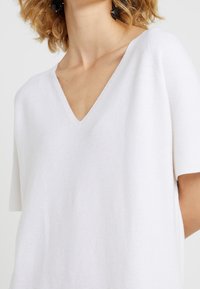 Haut tricoté blanc à manches courtes avec un décolleté en V, doté d'une texture lisse et d'une silhouette moderne. Design simple et minimaliste.