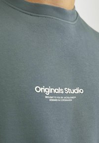 Sivi pamučni dukserica s rundastim izrezom, glatke teksture, i bijelim ispisanim natpisom "Originals Studio" te detaljima brenda.