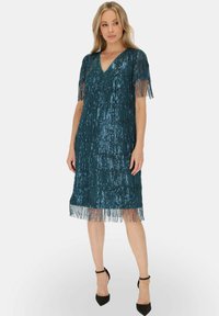 Miętowa sukienka z cekinami i frędzlami na rękawach oraz u dołu. Dekolt w kształcie litery V, długość do kolan, teksturowana tkanina, noszona z czarnymi obcasami.