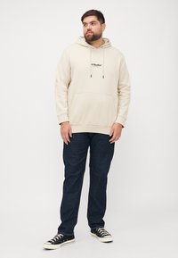 Cremefarbene Kapuze mit einer Fronttasche und Kordelzug. Kombiniert mit dunkelblauen Jeans und schwarzen Turnschuhen. Minimalistisches Design.