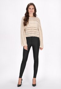Maglione beige lavorato a maglia con punti aperti e maniche lunghe, abbinato a pantaloni neri aderenti e tacchi alti neri.