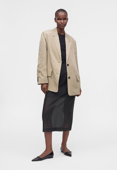 Γυναίκα με μπεζ oversized σακάκι, μαύρο διάφανο μεσαίο φόρεμα και μαύρα μυτερά φλατ παπούτσια, στέκεται μπροστά σε ένα απλό λευκό φόντο.
