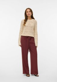 Femme debout portant un pull en tricot crème et un pantalon large taille haute bordeaux avec une ceinture à la taille, des talons ouverts noirs, sur un fond blanc.