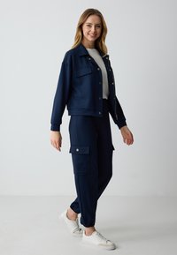 Navyblauwe jas met gouden hardware, voorzakken en een ontspannen pasvorm, gecombineerd met bijpassende cargobroek. Witte sneakers maken de outfit compleet.