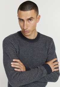 Marineblå strikket sweater med et struktureret mønster, rund hals og ribbede manchetter. Båret med armene krydset, hvilket viser hænder med lys hudtone.