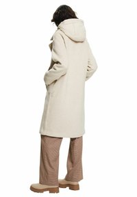 Abrigo largo beige con capucha, hecho de material suave, con bolsillos laterales y un corte recto, combinado con pantalones marrones con estampado y botas gruesas.