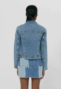 Lichtblauwe spijkerjasje met lange mouwen, knoopsluiting en achterzakken, gecombineerd met een patchwork spijkerrok in verschillende blauwtinten.