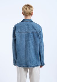 Giacca di jeans oversize in azzurro chiaro con collo classico, maniche lunghe e orlo grezzo. Presenta una spallina sul retro e una leggera texture.