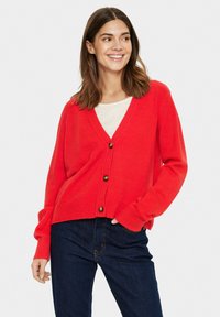 Röd kofta i stickat material med V-ringning, långa ärmar och träknappar. Matchad med mörkblå jeans.