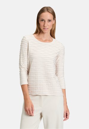Maglia beige a maniche lunghe con motivi ondulati testurizzati, scollatura rotonda e tessuto morbido. Abbinata a pantaloni chiari, design minimalista.