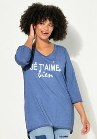 Camisa de algodón azul con cuello en V y mangas de tres cuartos, que presenta texto blanco y diseños de estrellas que dicen "JE T'AIME bien". Textura suave.