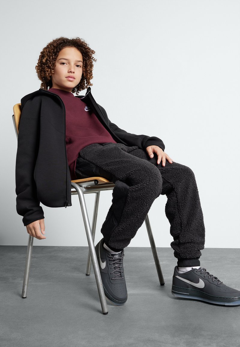 Un sweat à capuche zippé noir sur un sweat maroon, associé à un pantalon en polaire noir et des baskets grises Nike. Assis sur une chaise en métal.