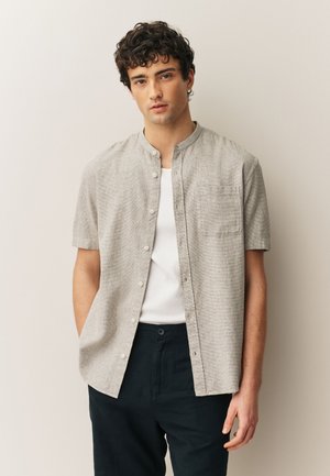 Jonge man met krullend donker haar, gekleed in een lichtgrijs kort mouw overhemd over een wit hemd en donkere broek, staand tegen een beige muur.