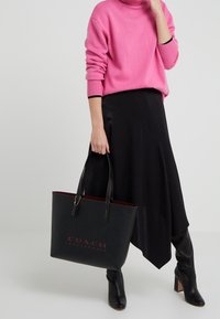 Czarna skórzana torba typu tote z czerwonym wnętrzem, z wytłoczonym logo "COACH LEATHERWARE", połączona z różowym swetrem i czarną spódnicą.