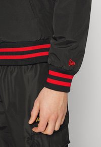 Svart nylonjacka med ribbade ärmslut och röda ränder samt en liten logotypapplikation. Tyget har en slät yta med en lätt lyster.