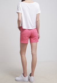 Vaaleanpunaiset puuvillashortsit, joissa on takataskut, yhdistettyinä valkoiseen lyhythihaiseen t-paitaan ja valkoisiin lenkkareihin, näkyvät takaa.