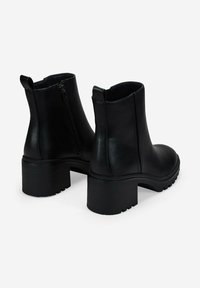 Un par de botas de piel negras hasta el tobillo con tacones de bloque, cremalleras laterales y tiradores sobre un fondo blanco.