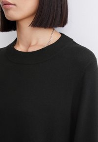 Pull noir en maille avec un col rond côtelé et des manches longues, présentant une texture douce. Collier chaîne argenté visible autour du cou.