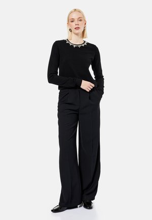 Blonde woman wearing a black long-sleeve top with jeweled neckline and wide-leg black trousers, standing with one hand in pocket.