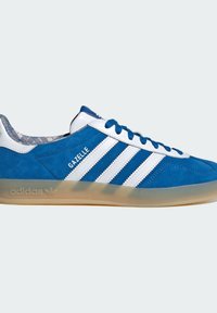 Baskets Adidas Gazelle bleues avec des bandes blanches, des lacets bleus et une semelle translucide sur un fond clair.