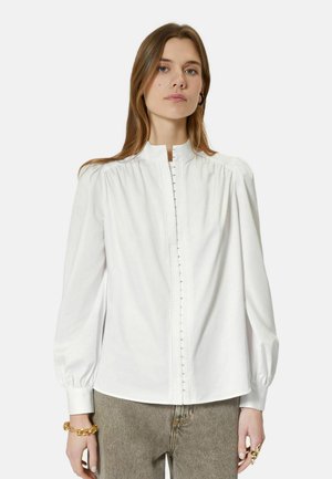 Donna che indossa una camicetta bianca a maniche lunghe con spalle plissettate e chiusure ad occhiello e gancio, abbinata a pantaloni grigio chiaro e gioielli in oro.