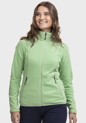 Frau lächelnd, trägt eine hellgrüne Fleecejacke mit Reißverschluss und zwei Reißverschlusstaschen sowie dunkle Hosen vor einem schlichten Hintergrund.