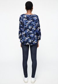 Námorníková květinová blůza s dlouhými rukávy, zdobená bílými květy a modrými listy. V páru s tmavými džíny a bílými teniskami. Zobrazený pohled zezadu.