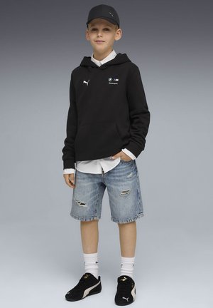 Dreng iført en sort Puma kasket og BMW Motorsport sort hættetrøje, hvid skjorte under, revne denimshorts, hvide sokker og sorte Puma sneakers.