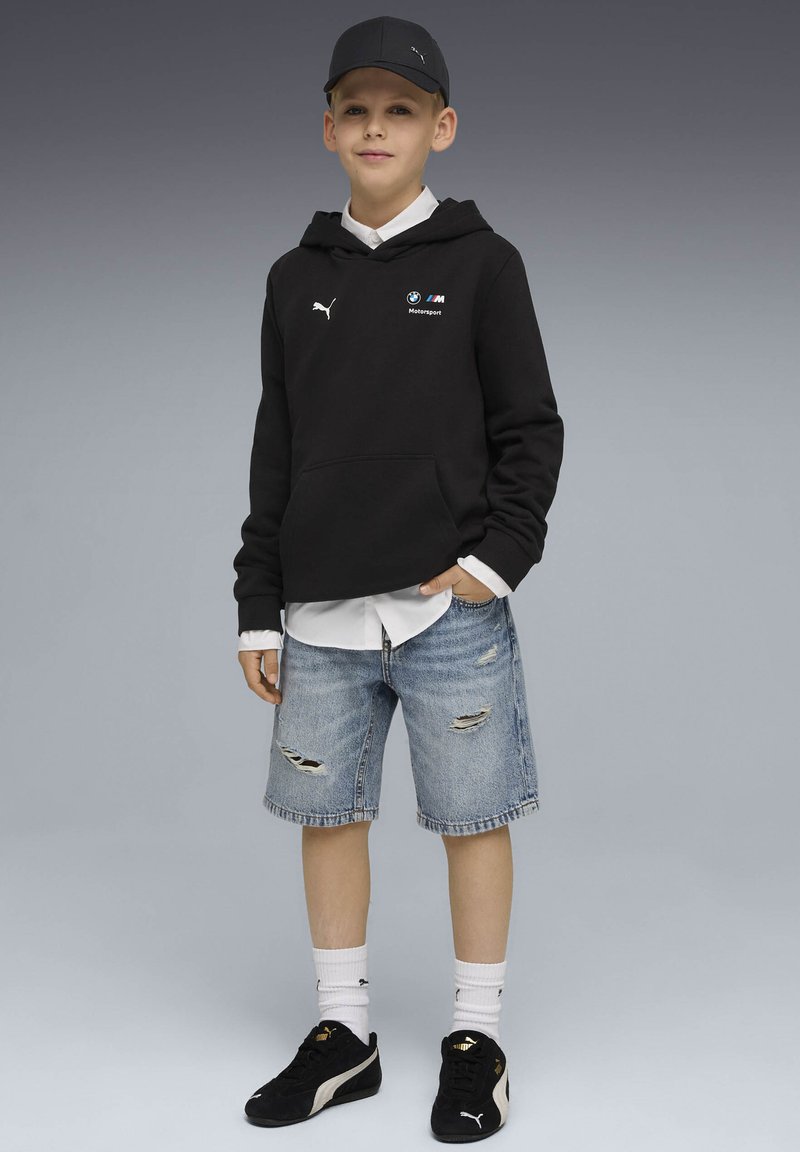 Garçon portant une casquette noire Puma et un sweat à capuche noir BMW Motorsport, un t-shirt blanc en dessous, un short en jean déchiré, des chaussettes blanches et des baskets noires Puma.