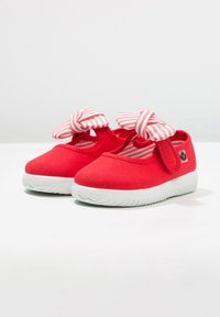 Coppia di scarpe rosse per bambini piccoli con suole in gomma bianche e fiocchi in tessuto a righe sulle fasce con velcro, mostrata su superficie bianca.