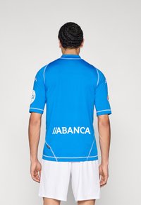 Modrý sportovní dres s krátkými rukávy, s bílými doplňky a logem "ABANCA" výrazně umístěným na zádech.