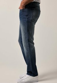 Jeans de mezclilla azul oscuro con corte ajustado, textura desgastada, cinco bolsillos y sutiles detalles de desgaste, combinados con zapatillas blancas.