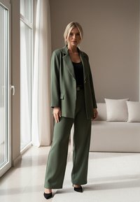 Oliwkowy garson z dopasowaną marynarką i szerokimi spodniami, połączony z czarną bluzką i szpilkami, na neutralnym tle.