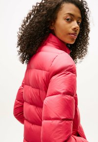 Czerwona kurtka puchowa z wysokim kołnierzem, z pikowanymi sekcjami i gładką, błyszczącą teksturą. Noszona z luźnymi, kręconymi włosami.