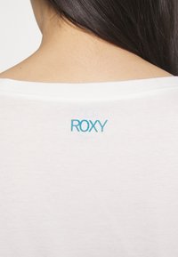 Valkoinen T-paita, jossa on pehmeä rakenne; siinä on pyöreä pääntie ja vaaleansininen "ROXY"-logo lähellä takakolttia.