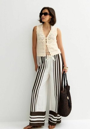 Mujer con top beige de crochet sin mangas, pantalones blancos de pierna ancha con rayas verticales marrones, sosteniendo un bolso tote oscuro tejido.