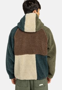 Fleece hoodie met een patchworkontwerp in donker groen, bruin, beige en teal; heeft een pluizige, gestructureerde oppervlakte en een grote capuchon.