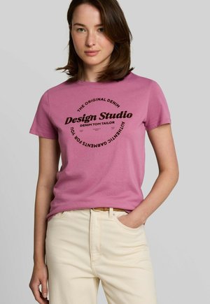 Femme aux cheveux bruns mi-longs, portant un t-shirt rose avec un texte noir et un pantalon beige clair, debout avec une main dans la poche.