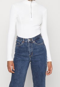 Wit geribd shirt met lange mouwen, een hoge kraag en zilveren ritssluiting, gecombineerd met donkerblauwe spijkerbroek met hoge taille.