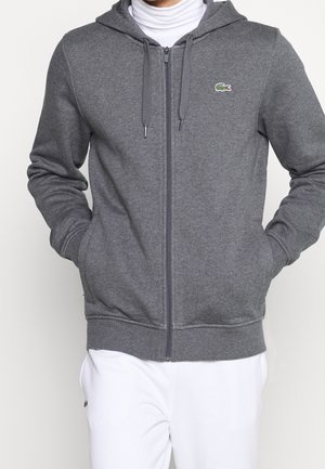 Persona que lleva una sudadera gris con capucha y cremallera, con cordones y un logo verde de cocodrilo, combinada con pantalones blancos.