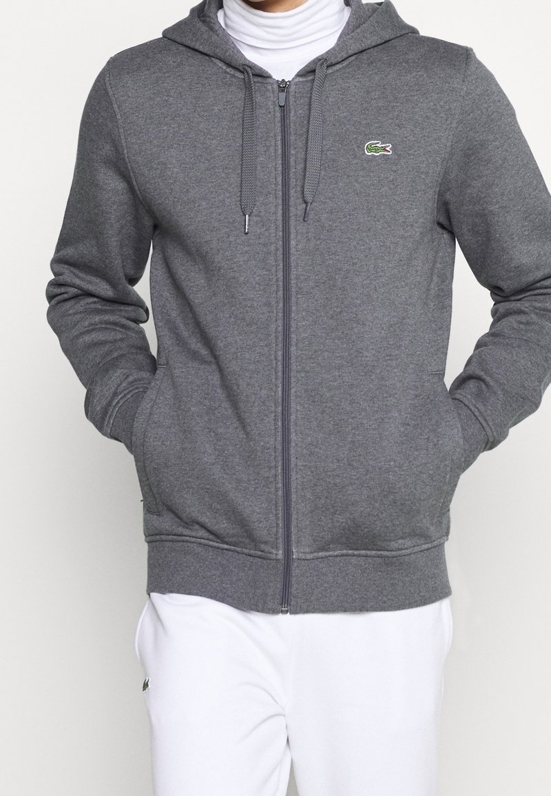 Personne portant un sweat à capuche gris zippé avec des cordons et un logo crocodile vert, associé à un pantalon blanc.