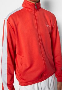 Chaqueta roja con cremallera hecha de tela suave. Presenta rayas laterales blancas, un cuello alto y detalles bordados. Incluye dos bolsillos frontales.