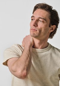 Beige T-shirt met korte mouwen en een ronde halslijn, gemaakt van textuurstof. Het model houdt zijn nek voorzichtig vast en toont een relaxte pasvorm.