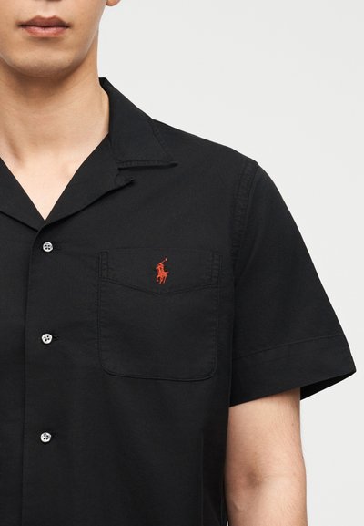 Άνδρας που φοράει μαύρο πουκάμισο με κοντά μανίκια και κουμπιά, με τσέπη στο στήθος και μικρό κόκκινο κεντητό λογότυπο αλόγου.