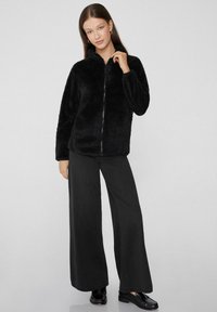 Czarna kurtka z sztucznego futra z zamkiem z przodu, wysokim kołnierzem, długimi rękawami i luźnym krojem. Połączona z szerokimi czarnymi spodniami i płaskimi butami.
