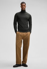 Ein Mann steht selbstbewusst da, trägt einen dunklen Rollkragenpullover, beiges Hosen und schwarze Schuhe, mit lässig in den Taschen versenkten Händen vor einem hellen Hintergrund.