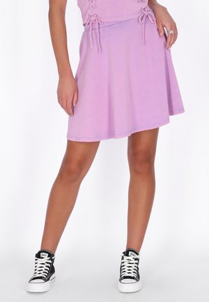 Kvinde iført lilla nederdel og matchende top med snøredetaljer, kombineret med sorte høje sneakers med hvide snørebånd og såler.